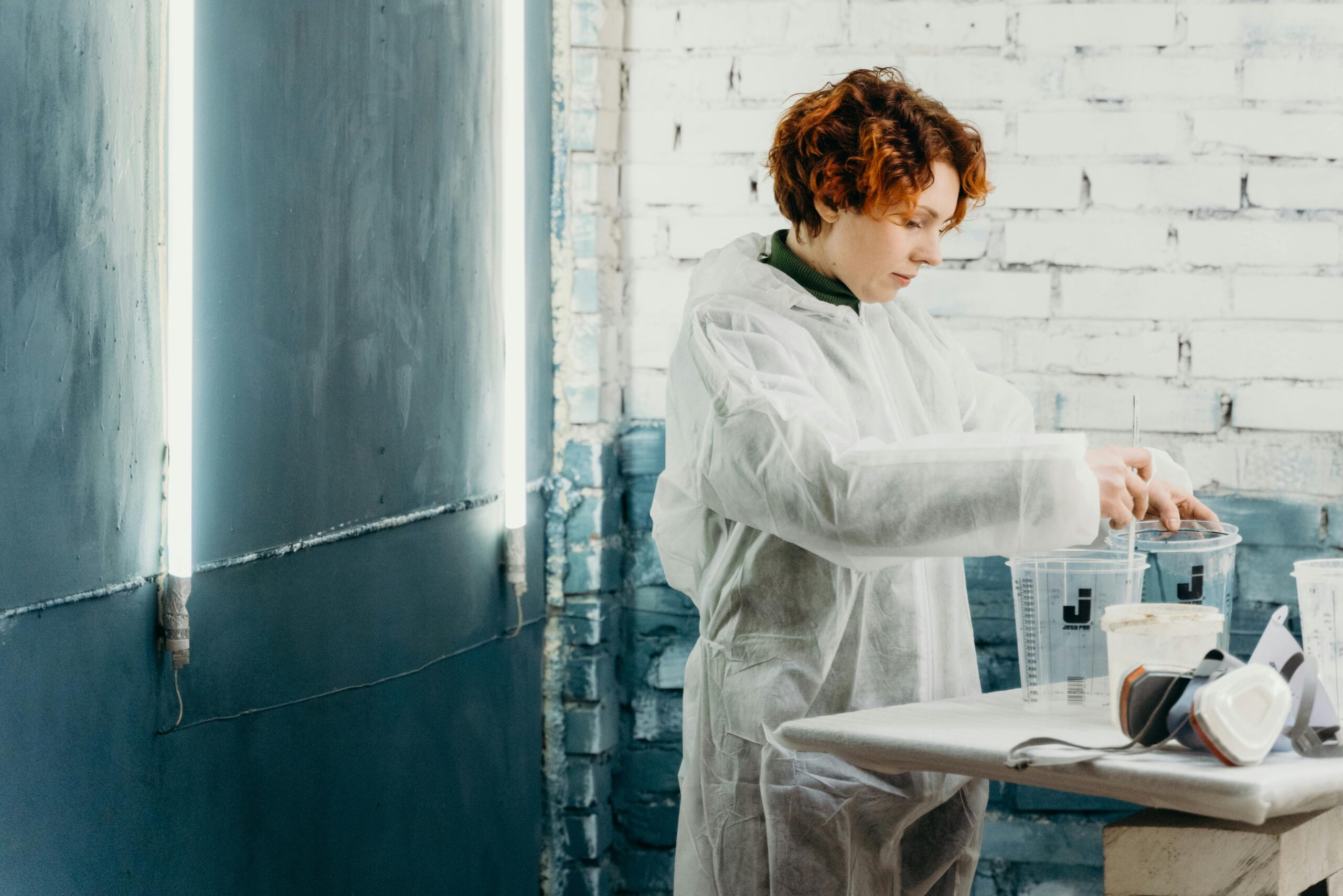 pexels-cottonbro-7493815 A woman wearing a white protective suit working in a studio environment with a rustic, painted brick wall and fluorescent lighting. She is stirring liquid in a measuring cup, with other cups and a respirator mask placed on a table nearby.